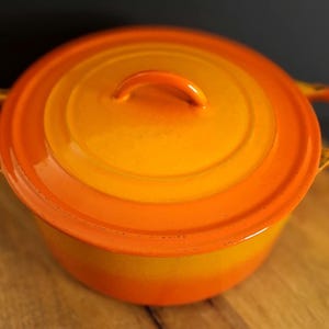 May include: A round, orange and yellow enameled cast iron Dutch oven with a matching lid. The pot features two side handles and a central handle on the lid. The exterior displays a gradient color transition from a bright orange rim to yellow.