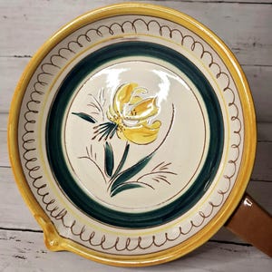 May include: A round, ceramic serving dish with a yellow rim and a green and white center. The center features a yellow flower design with green leaves. The dish has a brown handle and a small spout.