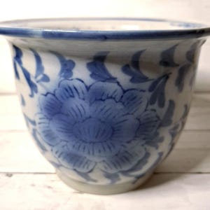 May include: A white and blue ceramic planter with a floral design. The planter features a large blue flower in the center and smaller floral accents around the rim. The rim is also trimmed with blue. The planter is a rounded shape.