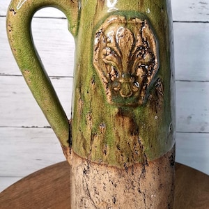 May include: A tall, green ceramic pitcher with a textured surface and a brown base. The pitcher features a raised fleur-de-lis emblem and a curved handle. The color is a mix of green and brown, with a rustic, aged appearance.