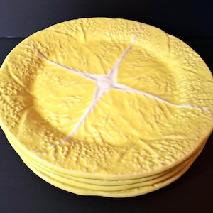 May include: A stack of round plates in a bright yellow color. Each plate features a textured surface that mimics the appearance of a cabbage leaf, complete with raised veins radiating from the center and a bumpy, organic leaf-like pattern.
