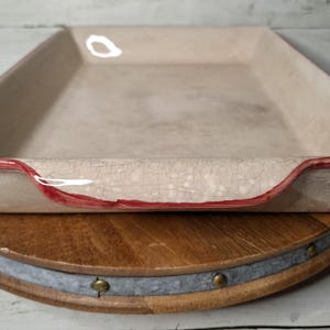 May include: A rectangular, off-white ceramic baking dish with a red rim, resting on a wooden serving board. The dish has a crackled surface and a slightly curved edge. The serving board is round and made of wood and metal.