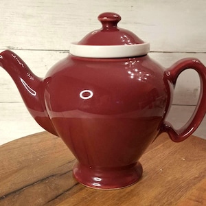 May include: A burgundy ceramic teapot with a white rim and lid. The teapot has a curved spout and a large handle. The teapot is sitting on a wooden surface.