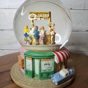 May include: A snow globe featuring a diner scene with miniature figures at a counter. The base includes a green cafe with the word "CAKE" and a blue convertible car. The globe is set on a round, tan base.