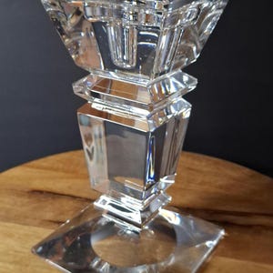 May include: A clear, faceted crystal candlestick holder with a square base and a geometric design. The candlestick has a tiered structure, with a square top to hold a candle. The object is on a wooden surface.