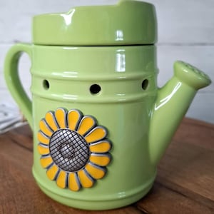 May include: A light green ceramic wax warmer shaped like a watering can. The front features a raised sunflower design with bright yellow petals and a textured gray center. The body includes a handle, a spout, and a series of small circular vents.
