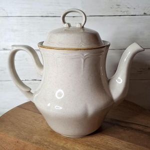May include: A beige ceramic teapot with a lid and handle. The teapot has a curved spout and a handle on the side. The lid has a loop handle. The teapot is sitting on a wooden surface.