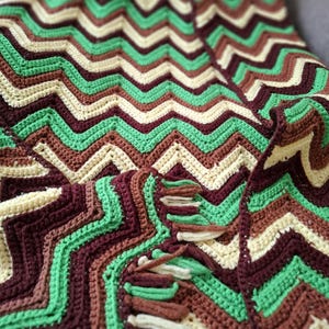 May include: A crocheted blanket featuring a repeating chevron pattern in alternating horizontal stripes of cream, light green, medium brown, and dark brown. The fabric has a textured stitch and includes short, knotted fringe along the edge.