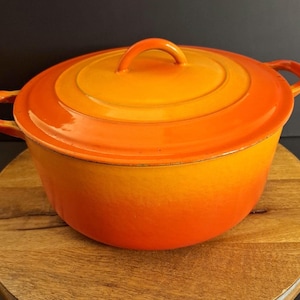 May include: A round enameled cast iron Dutch oven featuring a vibrant gradient color scheme that transitions from a bright yellow center on the lid to a deep orange hue along the base and sides, complete with two side handles and a matching lid.