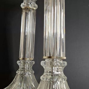 May include: A pair of clear glass candlesticks featuring a tall, fluted column design with a wide, ribbed base and a flared top. Each candlestick is accented with a thin metallic band at the neck and sits on a circular metallic base.