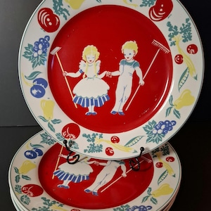 May include: A set of round ceramic plates featuring a bright red center with an illustration of two children holding hands and garden tools. The wide white rim is decorated with a colorful pattern of various fruits, vegetables, and green leaves.