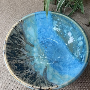 May include: A round ceramic bowl featuring a reactive glaze with a blend of vibrant blue, turquoise, and white tones. The interior surface displays a dramatic dark brown and black drip pattern that cascades from the rim towards the centre base.