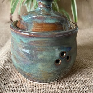 May include: A round ceramic jar with a matching lid, featuring a variegated glaze in shades of teal, blue, and earthy brown. The body of the jar includes a small cluster of three circular ventilation holes on the side, and a knob handle on the lid.