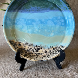 May include: A round ceramic plate featuring a multi-coloured glaze with horizontal bands of dark green, bright blue, and light beige. The bottom section displays a speckled, sandy texture with dark brown markings, resting on a black plastic stand.