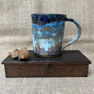 May include: A ceramic mug featuring a reactive glaze with layers of deep navy blue, bright sky blue, and earthy brown tones. The mug has a cylindrical body and a curved handle, showcasing a textured, mottled finish across the exterior surface.