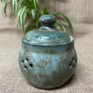 May include: A handmade ceramic jar with a lid, displaying a mottled teal and green glaze. The jar has a rounded form with several small, evenly spaced holes around the sides. The lid has a small knob on top.