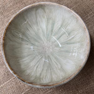 May include: A round ceramic bowl with a light green and white swirled pattern. The bowl has a brown rim and a glossy finish. The bowl is on a brown burlap surface.