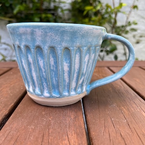 May include: A handmade ceramic mug with a light blue glaze and vertical ribbed design. The mug has a rounded base and a curved handle. The mug is sitting on a wooden surface.