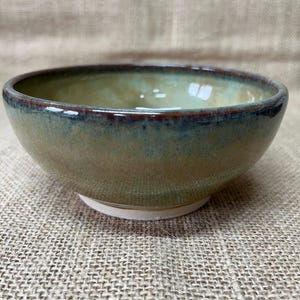 May include: A round ceramic bowl featuring a smooth, glossy glaze with blended tones of sage green, earthy tan, and deep brown along the rim. The bowl has a wide, open top and a circular base that is unglazed, revealing a light, matte off-white colour.