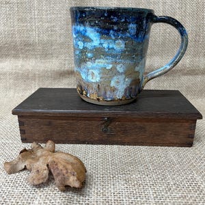 May include: A ceramic mug with a speckled glaze featuring a gradient of deep navy blue at the top transitioning into lighter shades of blue, white, and earthy brown tones. The mug has a curved handle and a cylindrical body with a textured finish.