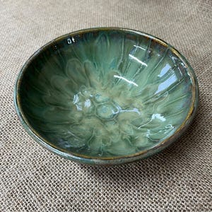 May include: A handcrafted ceramic bowl with a glossy, green glaze. The bowl has a rounded shape with a slightly raised rim and a textured interior. The glaze has a marbled effect, with shades of green and hints of brown.