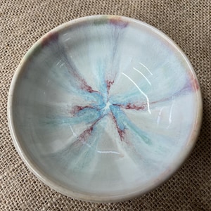 May include: A small, round ceramic bowl with a light blue and white interior design. The bowl's centre features a starburst pattern in shades of blue and maroon. The rim is a light beige colour.