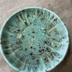 May include: A round ceramic bowl featuring a glossy glaze in shades of mint green and seafoam. The interior surface displays a radiating pattern of dark brown and black speckles that bleed outward from the centre toward the raised rim edge.