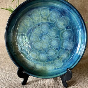 May include: A round ceramic bowl featuring a deep blue and teal glaze with a crystalline pattern. The interior surface displays a series of interconnected, light-coloured circular shapes that radiate from the centre, creating a distinct texture.