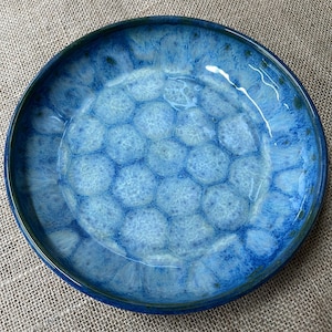 May include: A round ceramic plate with a blue glaze. The plate's centre has a honeycomb pattern, while the rim and outer edges are a darker shade of blue. The plate is a decorative piece.