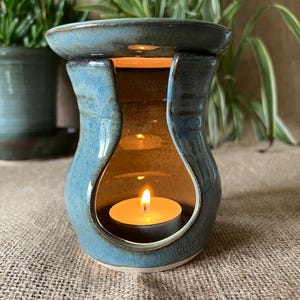 May include: A blue ceramic wax warmer with a glossy finish and a wide, arched opening. The piece features a flat top dish for wax melts and a hollow centre containing a small, lit white tea light candle that casts a warm glow from within.