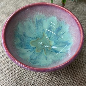 May include: A round ceramic bowl featuring a glossy glaze with a deep magenta rim that transitions into a vibrant turquoise and light green centre. The interior surface displays a starburst pattern of blue and green hues radiating from the base.