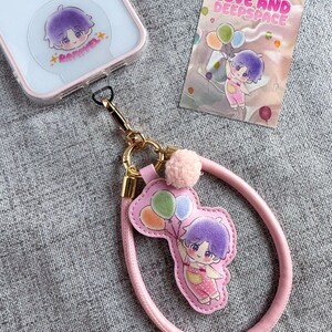 Op de afbeelding: Een roze telefoonriem met een stripfiguur die ballonnen vasthoudt. De riem is bevestigd aan een doorzichtige telefoonhoes met een bijpassende karaktersticker. Een kleine kaart met de tekst "LOVE AND DEEPSPACE" is ook zichtbaar.