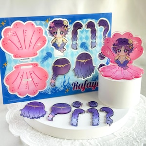 Könnte beinhalten: Ein Set aus Acryl-Anhängern und Aufklebern zum Thema Meerjungfrau. Die Anhänger zeigen eine Meerjungfrau-Figur mit lila Haaren und einer rosa Muschel. Die Aufkleber umfassen Muschelformen und Meerjungfrau-Haarteile. Das Set wird auf einer weißen Oberfläche präsentiert.