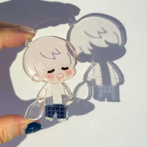 Op de afbeelding: Een heldere acryl bedel met een cartoonfiguur met gesloten ogen, roze wangen en een kleine tong. Het personage draagt een wit overhemd, een blauwe geruite broek en houdt een helder voorwerp vast. De bedel wordt omhoog gehouden en werpt een schaduw.