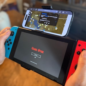 May include: A black Nintendo Switch game console with red and blue controllers, displaying a "Game Over" screen. A smartphone is mounted above, showing a map. The image is of a handheld gaming device.