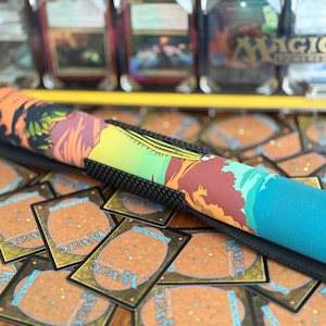 Op de afbeelding: Een opgerolde speelmat met een kleurrijk landschapsontwerp in oranje, groen en blauw. De mat ligt op een stapel Magic: The Gathering-kaarten en heeft een zwarte, getextureerde basis.
