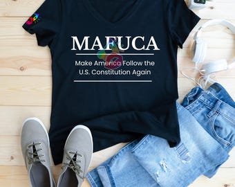 May include: Black V-neck t-shirt with the text "MAFUC*" and "Make America Follow the U.S. Constitution Again" in white. The shirt is paired with blue jeans, gray sneakers, and white headphones. A small plant and a rainbow graphic are also visible.