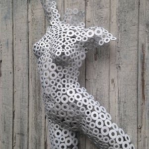 May include: A silver metal sculpture of a human torso, constructed from numerous circular washers. The sculpture is mounted on a weathered, light-colored wooden surface, creating a contrast between the industrial material and the rustic background.