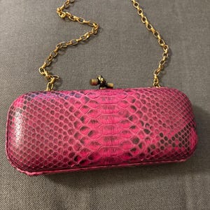 May include: A rectangular, hot pink clutch with a snakeskin pattern. The clutch has a gold-colored chain strap and a gold-colored clasp. The bag is made of a textured material.