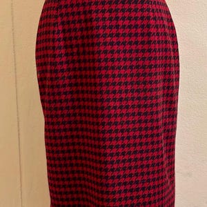 May include: A red and black houndstooth pattern skirt. The skirt is knee-length and has a classic, tailored design. The fabric has a woven texture, and the pattern is a repeating geometric design. The skirt is displayed on a mannequin.