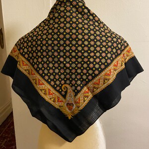 May include: A square black scarf with a repeating floral pattern in yellow, orange, and red. The border features a geometric design and a paisley motif. The scarf is displayed on a mannequin.