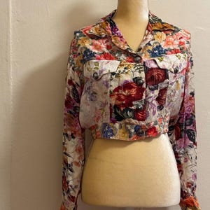 May include: A cropped long-sleeve jacket featuring a vibrant floral pattern with red, pink, yellow, and blue flowers on a light background. The jacket includes a pointed collar, two chest pockets with flaps, and silver-toned metal buttons.