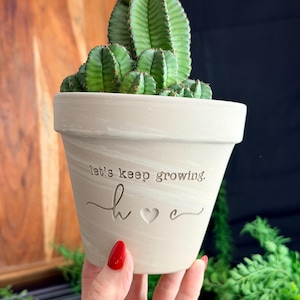 May include: A light-colored ceramic flower pot with a green cactus. The pot has the words "let's keep growing. h ♡ e" etched on the side. The cactus has a ribbed texture and is a vibrant green color.
