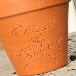 May include: A terracotta-colored flower pot with the engraved text "The Hendersons" in a decorative script. The pot features ornate, swirling embellishments above and below the name, adding a touch of elegance to the design. The pot is sitting on a decorative metal table.