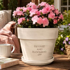 May include: A large, off-white ceramic flower pot with the words "Retired and Blooming" in dark gray lettering. The pot is filled with vibrant pink azalea flowers. A saucer is beneath the pot. A book and a mug are on the table.