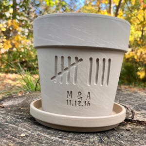 May include: A light beige ceramic flower pot with a matching saucer. The pot has vertical cut-out lines and a carved tally mark design. The initials "M & A" and the date "11.12.16" are carved below the tally marks. The pot is sitting on a wooden surface.