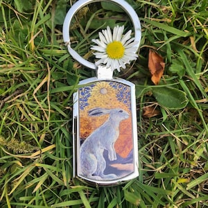 Puede incluir: Un llavero plateado con una margarita blanca unida al anillo. El dije del llavero presenta una ilustración colorida de un conejo blanco en un campo con un sol y un cielo azul.