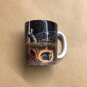 May include: A black ceramic mug with a white handle. The mug features a colorful illustration of badgers in a forest setting at night. The illustration includes a starry sky, a tree, and a badger family.