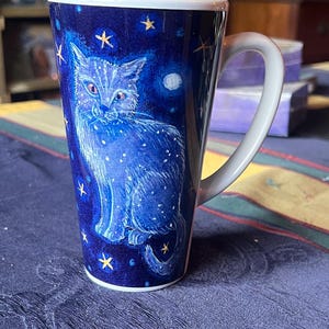 May include: A white ceramic mug with a blue cat design. The cat is surrounded by gold stars on a dark blue background. The mug has a white handle.