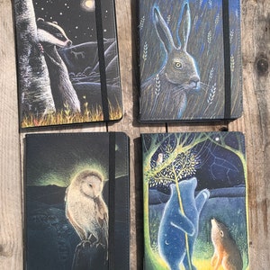 May include: Four black journals with different animal illustrations. The covers feature a badger, a hare, an owl, and a bear with a wolf. Each journal has a black elastic closure.